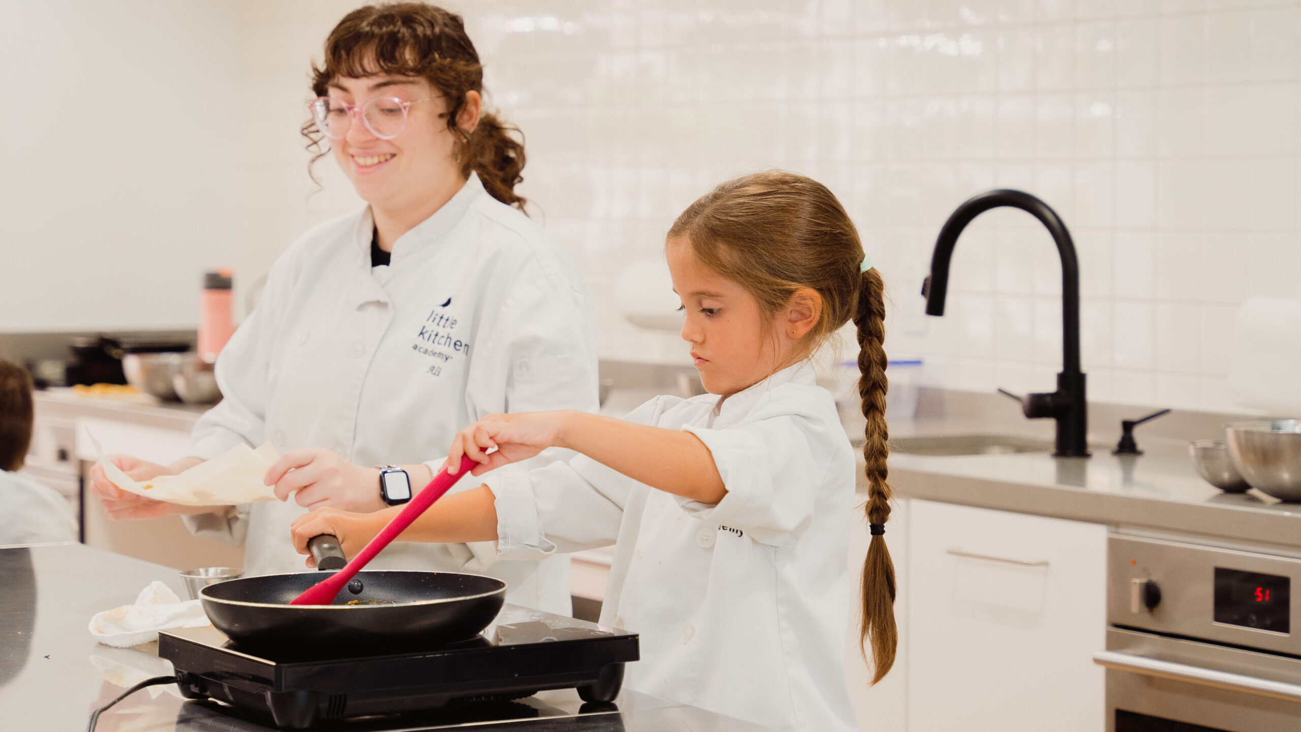 A student at Little Kitchen Academy uses a spatula to cook at a stovetop under the guidance of an instructor. Both are wearing white chef coats in a clean, modern kitchen setting. This Montessori-inspired culinary environment empowers students to safely practice real-life cooking skills, fostering independence, focus, and confidence with supportive mentorship.